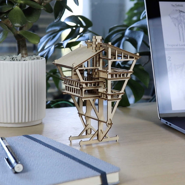 De Tropical Lookout DIY houten modelbouwkit van Tiny Treehouses Europe staat op een bureau naast een gesloten notitieboekje met een pen, een potplant en een open laptop met instructies.