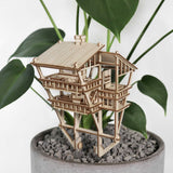 De Tropical Lookout van Tiny Treehouses Europe is een klein doe-het-zelf houten modelbouwpakket van een huis met meerdere verdiepingen tussen groene bladeren en grijze kiezels in een lichte ronde plantenbak, die de sfeer creëert van een gezellig junglehuis.