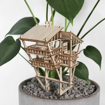 De Tropical Lookout van Tiny Treehouses Europe is een klein doe-het-zelf houten modelbouwpakket van een huis met meerdere verdiepingen tussen groene bladeren en grijze kiezels in een lichte ronde plantenbak, die de sfeer creëert van een gezellig junglehuis.
