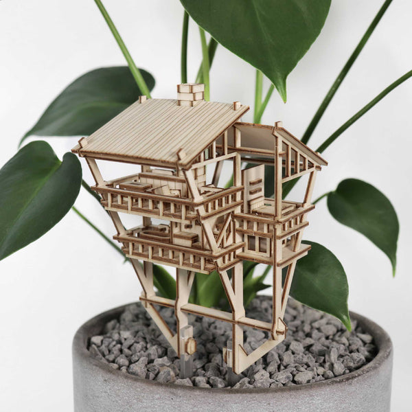 De Tropical Lookout van Tiny Treehouses Europe is een klein doe-het-zelf houten modelbouwpakket van een huis met meerdere verdiepingen tussen groene bladeren en grijze kiezels in een lichte ronde plantenbak, die de sfeer creëert van een gezellig junglehuis.