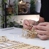Handen van een persoon zetten de Tropical Lookout DIY houten bouwset van Tiny Treehouses Europe in elkaar op een witte tafel met plantendecor en modelstukken. Zachte LED-verlichting draagt bij aan de gezellige, op de jungle geïnspireerde sfeer van deze miniatuur boomhut.