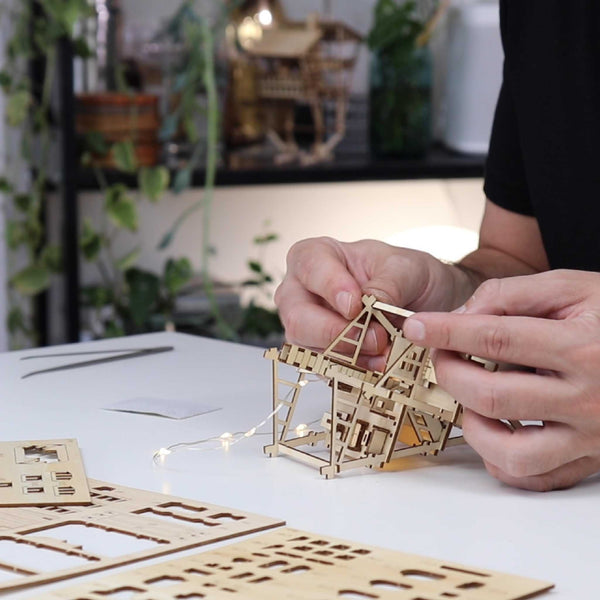 Handen van een persoon zetten de Tropical Lookout DIY houten bouwset van Tiny Treehouses Europe in elkaar op een witte tafel met plantendecor en modelstukken. Zachte LED-verlichting draagt bij aan de gezellige, op de jungle geïnspireerde sfeer van deze miniatuur boomhut.