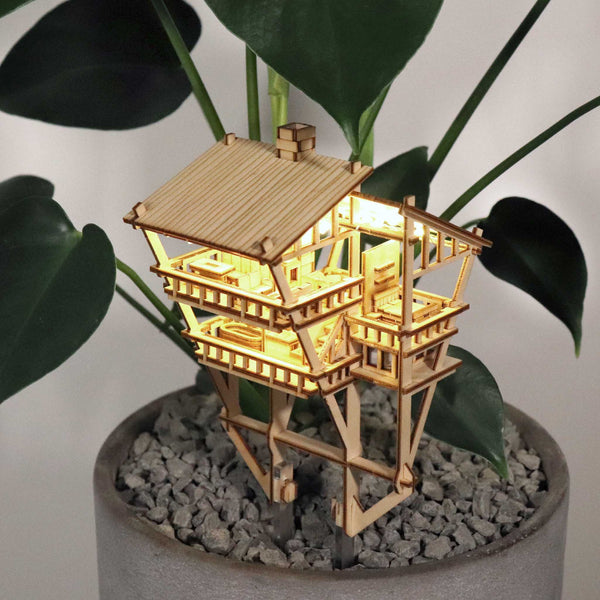 De Tropical Lookout van Tiny Treehouses Europe is een klein, verlicht houten bouwpakket van een junglehuis met twee verdiepingen. Het staat bovenop grind in een potplant, gloeit met LED-lampjes en wordt omringd door weelderige groene bladeren voor een gezellige, grillige touch.