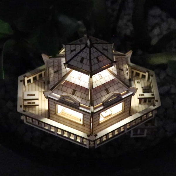 De Woodland Outpost van Tiny Treehouses Europe is een klein, verlicht doe-het-zelf houten modelbouwpakket van een zeshoekig huis met gedetailleerde leuningen en ramen, van bovenaf gezien tegen een donkere, kiezelachtige achtergrond.