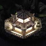 De Woodland Outpost van Tiny Treehouses Europe is een klein, verlicht doe-het-zelf houten modelbouwpakket van een zeshoekig huis met gedetailleerde leuningen en ramen, van bovenaf gezien tegen een donkere, kiezelachtige achtergrond.