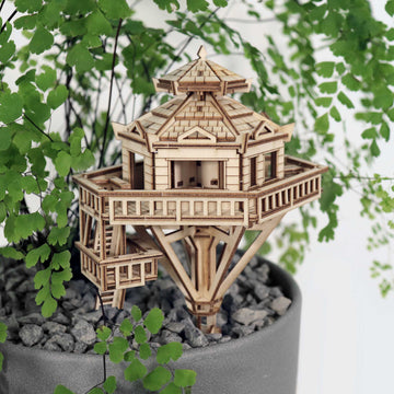 De Woodland Outpost van Tiny Treehouses Europe is een charmant zeshoekig doe-het-zelf houten modelbouwpakket, zittend op grijze kiezels in een plantenpot en omringd door delicate varenbladeren - perfect om speelsheid toe te voegen aan elke ruimte.