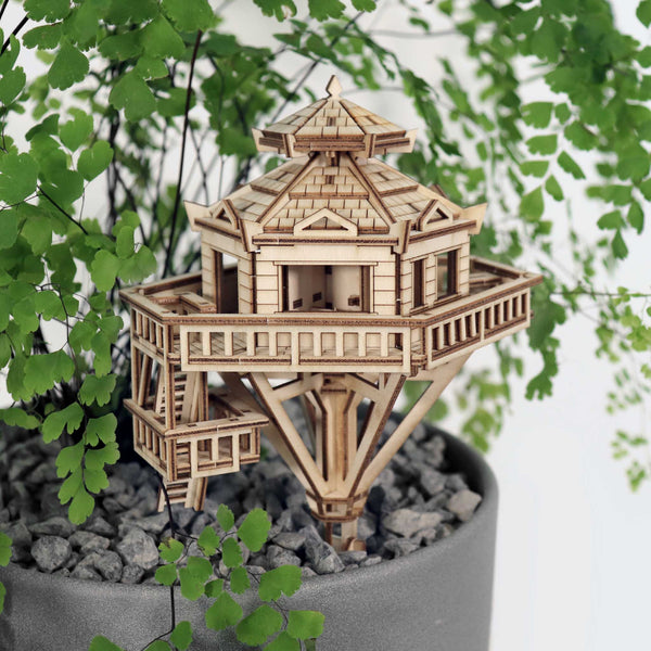 De Woodland Outpost van Tiny Treehouses Europe is een charmant zeshoekig doe-het-zelf houten modelbouwpakket, zittend op grijze kiezels in een plantenpot en omringd door delicate varenbladeren - perfect om speelsheid toe te voegen aan elke ruimte.