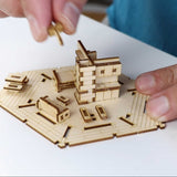 Een persoon zet Tiny Treehouses Europe's Woodland Outpost, een modern houten modelbouwpakket met meerdere verdiepingen, in elkaar door een stuk bovenop te plaatsen terwijl houten miniatuurmeubels en accessoires aan de basis worden geplaatst.