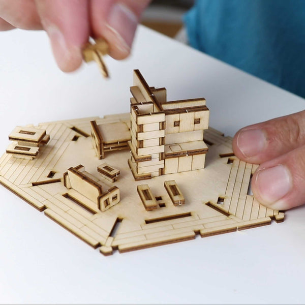 Een persoon zet Tiny Treehouses Europe's Woodland Outpost, een modern houten modelbouwpakket met meerdere verdiepingen, in elkaar door een stuk bovenop te plaatsen terwijl houten miniatuurmeubels en accessoires aan de basis worden geplaatst.