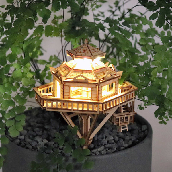 De Woodland Outpost van Tiny Treehouses Europe is een doe-het-zelf houten modelbouwpakket met LED-verlichting, ontworpen om op een plantenpot te staan gevuld met kleine grijze steentjes en omringd door fijne groene blaadjes.