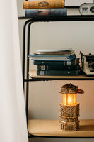 Op een houten plank liggen gestapelde boeken, losse papieren, een oude typemachine en de WoodTrick Antique Lantern - een houten bouwpakket van WoodTrick in de vorm van een gotisch gebouw, zacht gloeiend met warm licht.