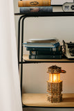 Op een houten plank liggen gestapelde boeken, losse papieren, een oude typemachine en de WoodTrick Antique Lantern - een houten bouwpakket van WoodTrick in de vorm van een gotisch gebouw, zacht gloeiend met warm licht.