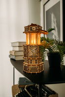 De WoodTrick Antique Lantern, een houten bouwpakket van WoodTrick met LED-verlichting en sierlijke uitsnijdingen, straalt warm op een zwarte plank tussen boeken, een potplant en kunst in een sfeervol verlichte kamer.