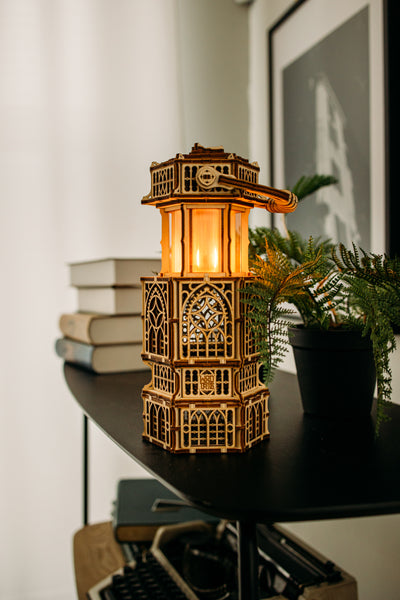 De WoodTrick Antique Lantern, een houten bouwpakket van WoodTrick met LED-verlichting en sierlijke uitsnijdingen, straalt warm op een zwarte plank tussen boeken, een potplant en kunst in een sfeervol verlichte kamer.
