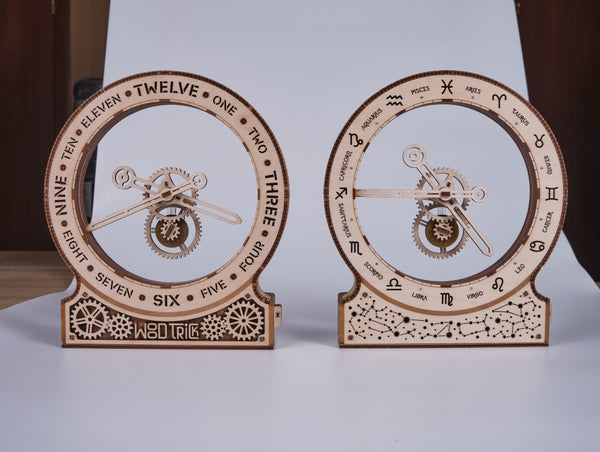 Twee WoodTrick Kinetische Klok: Modern - Houten Klok Bouwpakket worden naast elkaar getoond. Op de linker staan tijdlabels, op de rechter dierenriemsymbolen. Beide klokken met zichtbare tandwielen hebben gedetailleerde uitgesneden ontwerpen van WoodTrick.