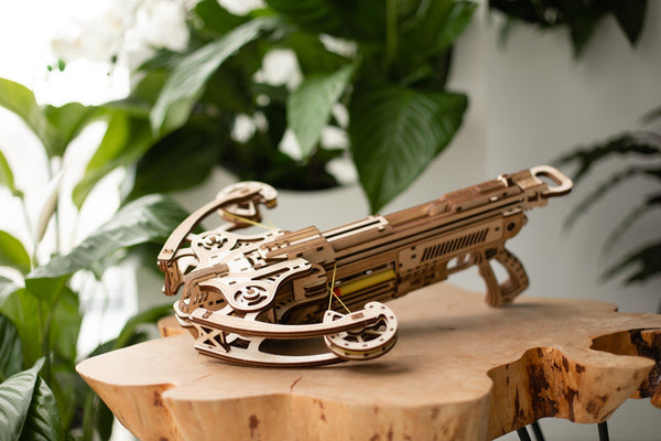 De WoodTrick Bullet Boltcaster - Houten Kruisboog Bouwpakket van WoodTrick wordt tentoongesteld op een tafel van natuurlijk hout, omringd door groene planten en witte bloemen, die het werkend schietmechanisme benadrukken.