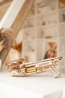 De WoodTrick Bullet Boltcaster, een houten kruisboog bouwpakket voor volwassenen van het merk WoodTrick, staat op een witte ondergrond in een lichte kamer met planken en neutrale decoratie op de achtergrond.
