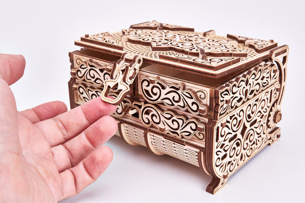 Een hand opent een WoodTrick Schatkist - Sieradendoos Bouwpakket met Kristallen, waarbij het ingewikkelde lasergesneden ontwerp en de kristallen accenten opvallen. Het decoratieve slot en de elegante afwerking steken af tegen de effen witte achtergrond. Merk: WoodTrick.