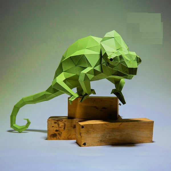 De Kameleon op Tak 3D Papier Model van SlimSpul nederland b.v. is een groene, geometrische kameleon sculptuur in origami-stijl, weergegeven op houten blokken tegen een effen achtergrond.