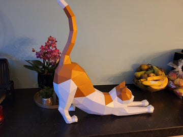 Het SlimSpul nederland b.v. Rekkende Kat 3D Papier Model - Elegant Huisdier Bouwpakket, een geometrische oranje en witte DIY sculptuur, staat op een aanrecht in de buurt van een roze orchidee, dienblad, fruitschaal en appels.