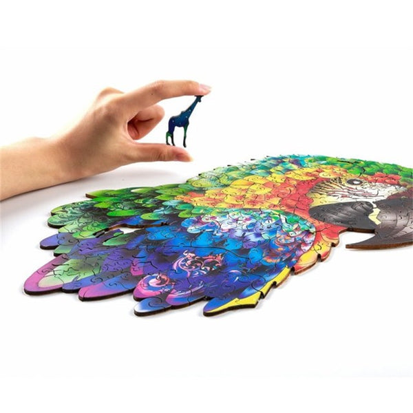 Een hand legt een blauw stukje in de vorm van een giraffe op de Papegaai Houten Puzzel (A3 formaat) - Kleurrijk Vogel Figuurpuzzel van SlimSpul nederland b.v., waardoor een kleurrijke vogelpuzzel ontstaat op een wit vlak.