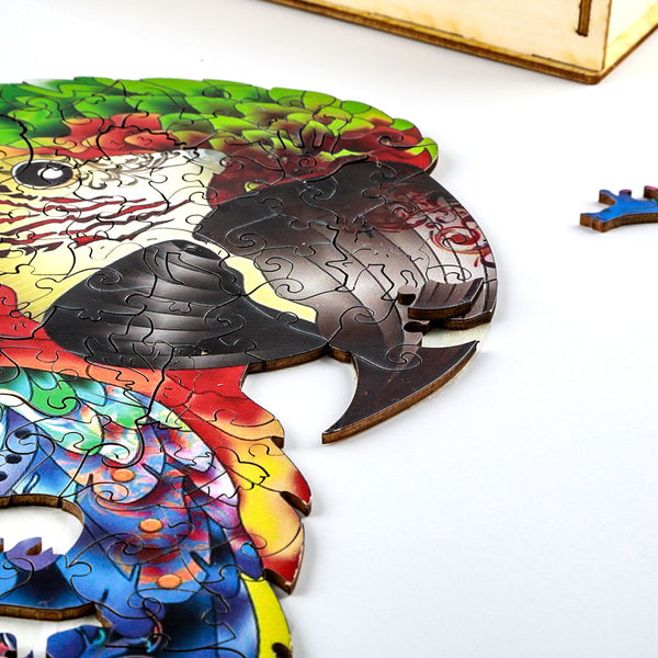 Een gedeeltelijk voltooide SlimSpul nederland b.v. Papegaai Houten Puzzel (A3 formaat) - Kleurrijk Vogel Figuurpuzzel, waarvan een stukje bij de snavel ontbreekt, toont levendige kleuren en een extra stukje dat op een wit vlak rust.