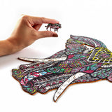 Een hand houdt een Grijze Olifant Houten Puzzel (A3 formaat) - Majestueus Dier Figuurpuzzel van SlimSpul nederland b.v. vast, met levendige, wervelende ontwerpen - ideaal voor beenpuzzel volwassenen die op zoek zijn naar een unieke uitdaging.