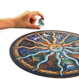 Een hand houdt een puzzelstukje vast boven de SlimSpul nederland b.v. Dierenriem Houten Puzzel (A4+ of A3), een astrologische figuurpuzzel met kleurrijke dierenriemsymbolen op een witte achtergrond.