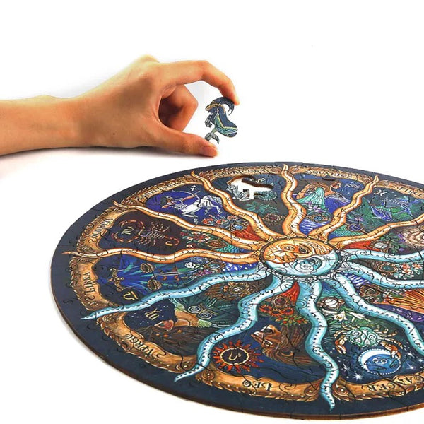 Een hand houdt een puzzelstukje vast boven de SlimSpul nederland b.v. Dierenriem Houten Puzzel (A4+ of A3), een astrologische figuurpuzzel met kleurrijke dierenriemsymbolen op een witte achtergrond.