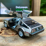 Het DeLorean Tijdmachine Metalen Bouwpakket | Back to the Future Auto van SlimSpul nederland b.v. heeft een open vleugeldeur ontwerp met een zilveren afwerking en gedetailleerd interieur, weergegeven op hout tegen een groene achtergrond.