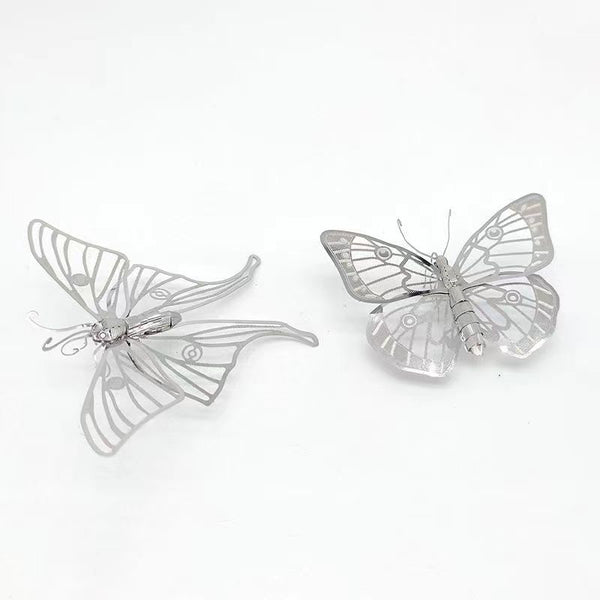 Het Vlinders Metalen Bouwpakket (Set van 2) van SlimSpul nederland b.v. heeft delicate metalen vlindermodellen in metalen modelbouwstijl, met transparante vleugels - één open, één gedeeltelijk gesloten - op een witte achtergrond.