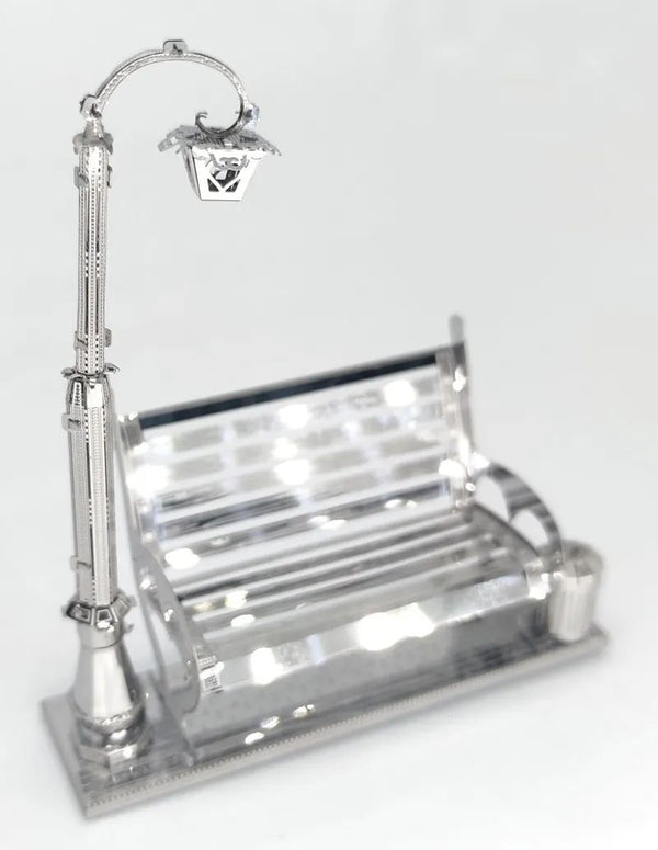 Het Parkbankje met Lantaarn Metalen Bouwpakket - Miniatuur Stadsgezicht van SlimSpul nederland b.v. heeft een gedetailleerde zilveren en glazen parkbank, straatlantaarn en afvalbak op een rechthoekige voet, perfect voor miniatuur stadsgezichten.