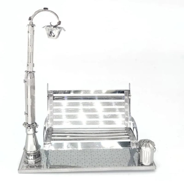Het Parkbankje met Lantaarn Metalen Bouwpakket - Miniatuur Stadsgezicht van SlimSpul nederland b.v. is een klein metalen model met een parkbank, straatlantaarn en prullenbak op een betegelde voet, met reflecterende oppervlakken en fijne details.