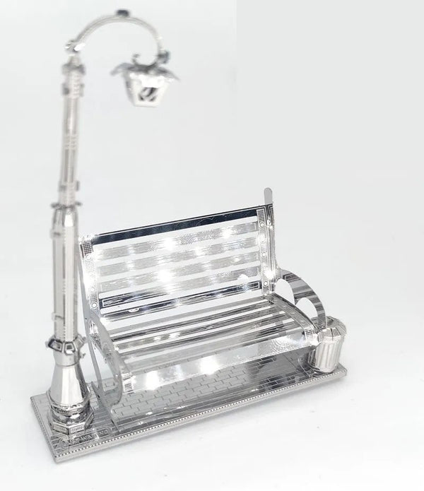 Het Parkbankje met Lantaarn Metalen Bouwpakket van SlimSpul nederland b.v. is een miniatuur stadsgezicht met een glanzend zilveren bankje, straatlantaarn en afvalbak op een gestructureerde voet, perfect voor verzamelaars en modelbouwliefhebbers.
