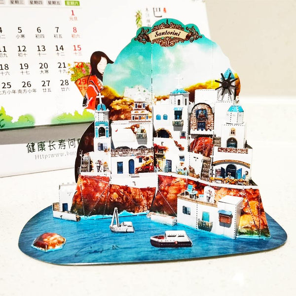 Een Stadsgezichten Metalen Bouwpakket (Diverse Steden) van SlimSpul nederland b.v. -een architectonische miniatuur van Santorini met witte gebouwen, blauwe koepels en boten- getoond voor een kalender op een wit oppervlak.