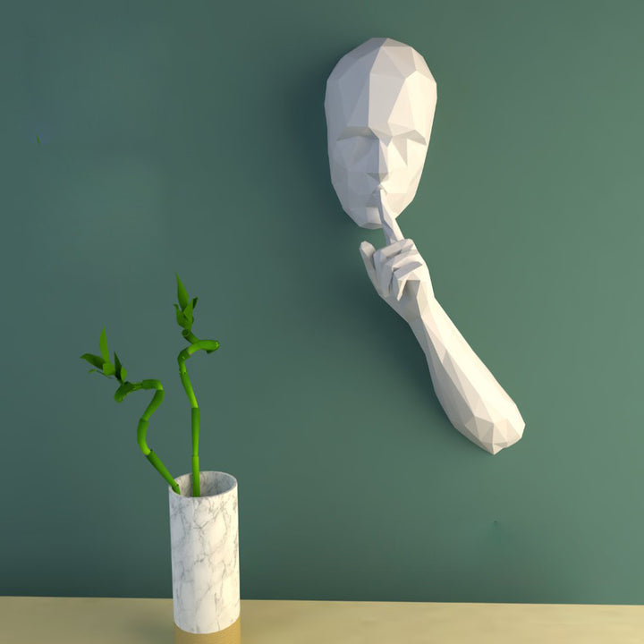 Wit low-poly Sssssssttt-papiermodel met gezicht, hand en vinger voor de lippen tegen een groene wand