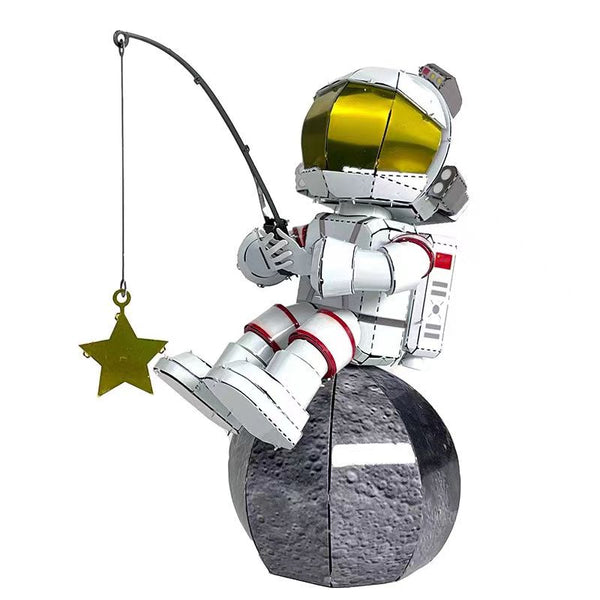The Astronaut Vissend naar Sterren Metalen Bouwpakket van SlimSpul nederland b.v. toont een cartoon ruimtevaarder in wit-rood pak met gouden vizier, zittend op een maan en vissend naar een gouden ster-perfect voor decoratie heelal liefhebbers.