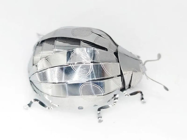 Het Lieveheersbeestje Metalen Bouwpakket van SlimSpul nederland b.v. is een glanzend, metalen lieveheersbeestje met een gesegmenteerd lichaam en poten, voorzien van gedetailleerde, reflecterende texturen en weergegeven op een witte achtergrond.