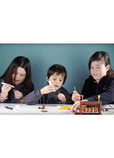 Drie kinderen zitten aan een tafel te knutselen met boetseerklei en gereedschap. Onder hen is het Rups Houtconstructie Bouwpakket - Bewegend Insect Model van SlimSpul nederland b.v., dat creativiteit stimuleert met hands-on bouwplezier.