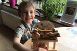 Een jongetje glimlacht tijdens het bouwen van de Aardman Sleepy Farmer Houtconstructie - Beginner Model (Timberkits) van SlimSpul nederland b.v. aan een tafel bij een raam, met een groene potplant en papieren op de achtergrond - ideaal voor de beginnende modelbouw.