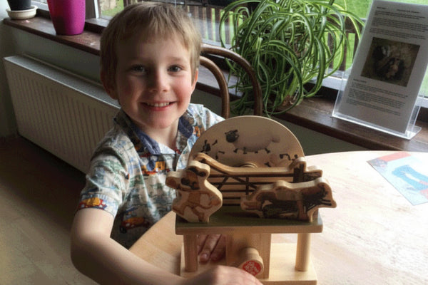 Een jongetje glimlacht tijdens het bouwen van de Aardman Sleepy Farmer Houtconstructie - Beginner Model (Timberkits) van SlimSpul nederland b.v. aan een tafel bij een raam, met een groene potplant en papieren op de achtergrond - ideaal voor de beginnende modelbouw.