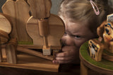 Een jong kind met een roze haarspeld bekijkt en speelt aandachtig met de Aardman Jumping Timmy Houtconstructie - Beginner Model (Timberkits) van SlimSpul nederland b.v., terwijl er ook houten Shaun het Schaap figuren op tafel staan.