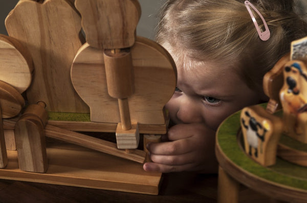 Een jong kind met een roze haarspeld bekijkt en speelt aandachtig met de Aardman Jumping Timmy Houtconstructie - Beginner Model (Timberkits) van SlimSpul nederland b.v., terwijl er ook houten Shaun het Schaap figuren op tafel staan.
