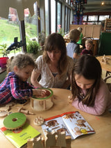 Een vrouw en twee jonge meisjes zetten een SlimSpul nederland b.v. Aardman Flock Roundabout Houtconstructie - Draaimolen Model (Timberkits Beginner) in elkaar aan een tafel; één bestudeert de instructies terwijl de anderen spelen bij grote ramen.